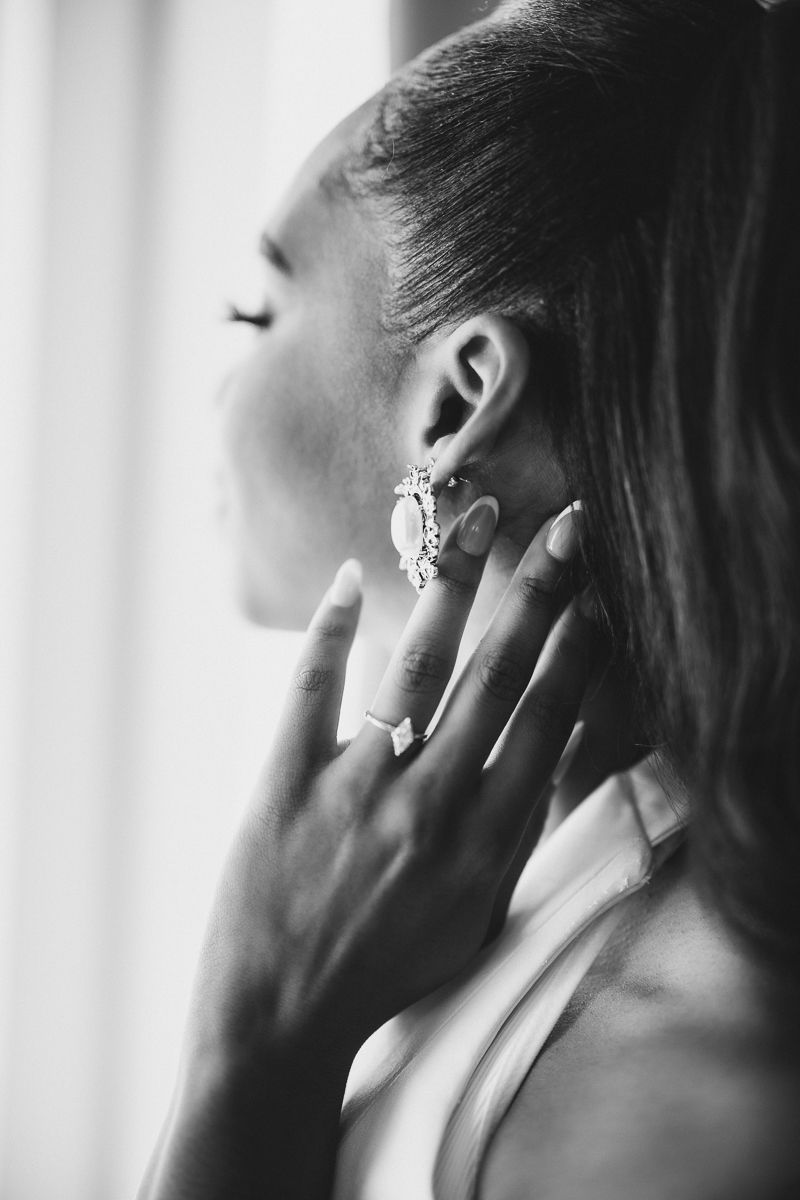 Black and white photo of a bride touching her elegant crystal earring and diamond ring near a window.