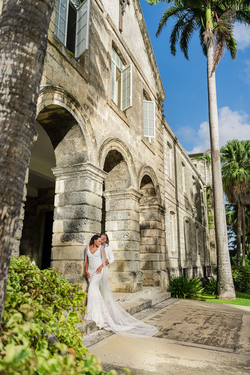 A couple in wedding attire poses along an arched stone walkway with tropical palm trees and blue sky in the background.