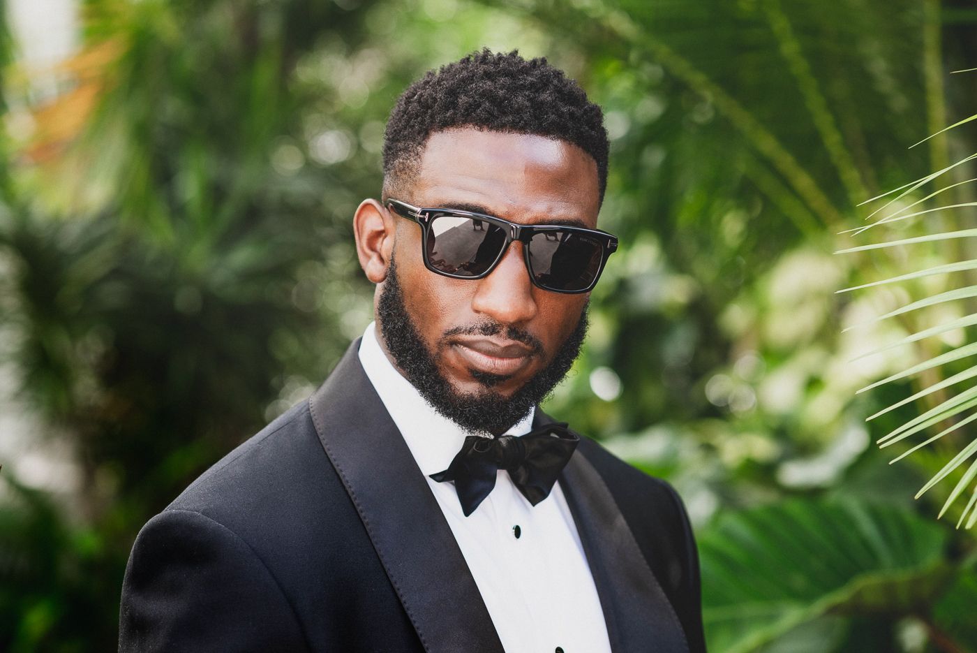 Formal portrait with sunglasses and bow tie against tropical foliage background.