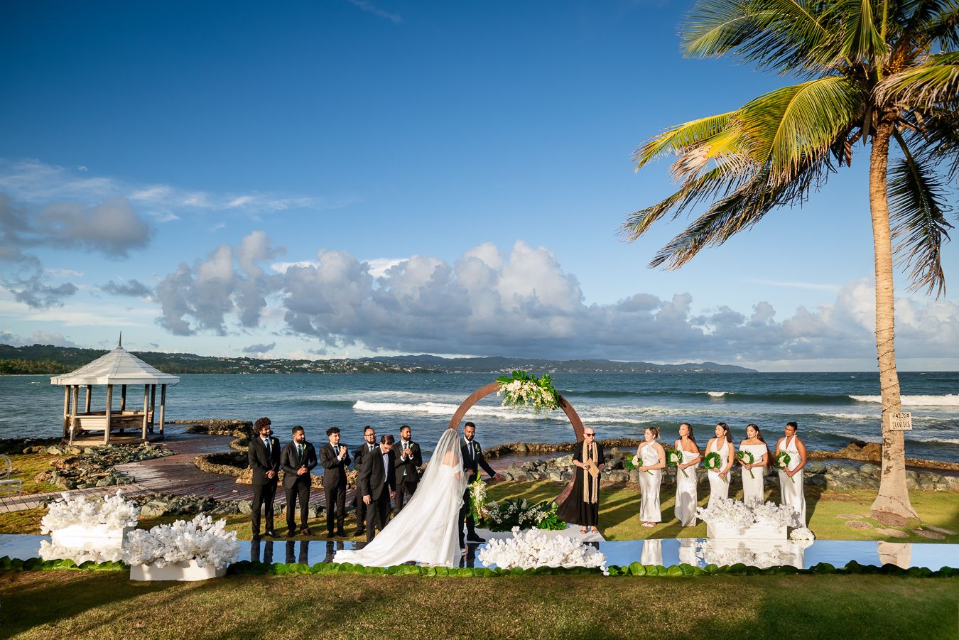 Wedding party lines up on oceanfront lawn with palm trees and gazebo for ceremony photos.