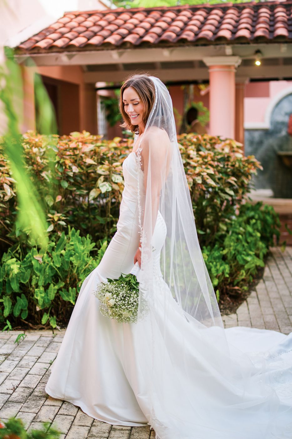 A bride poses in an elegant white gown with long veil near Spanish-style architecture and gardens.