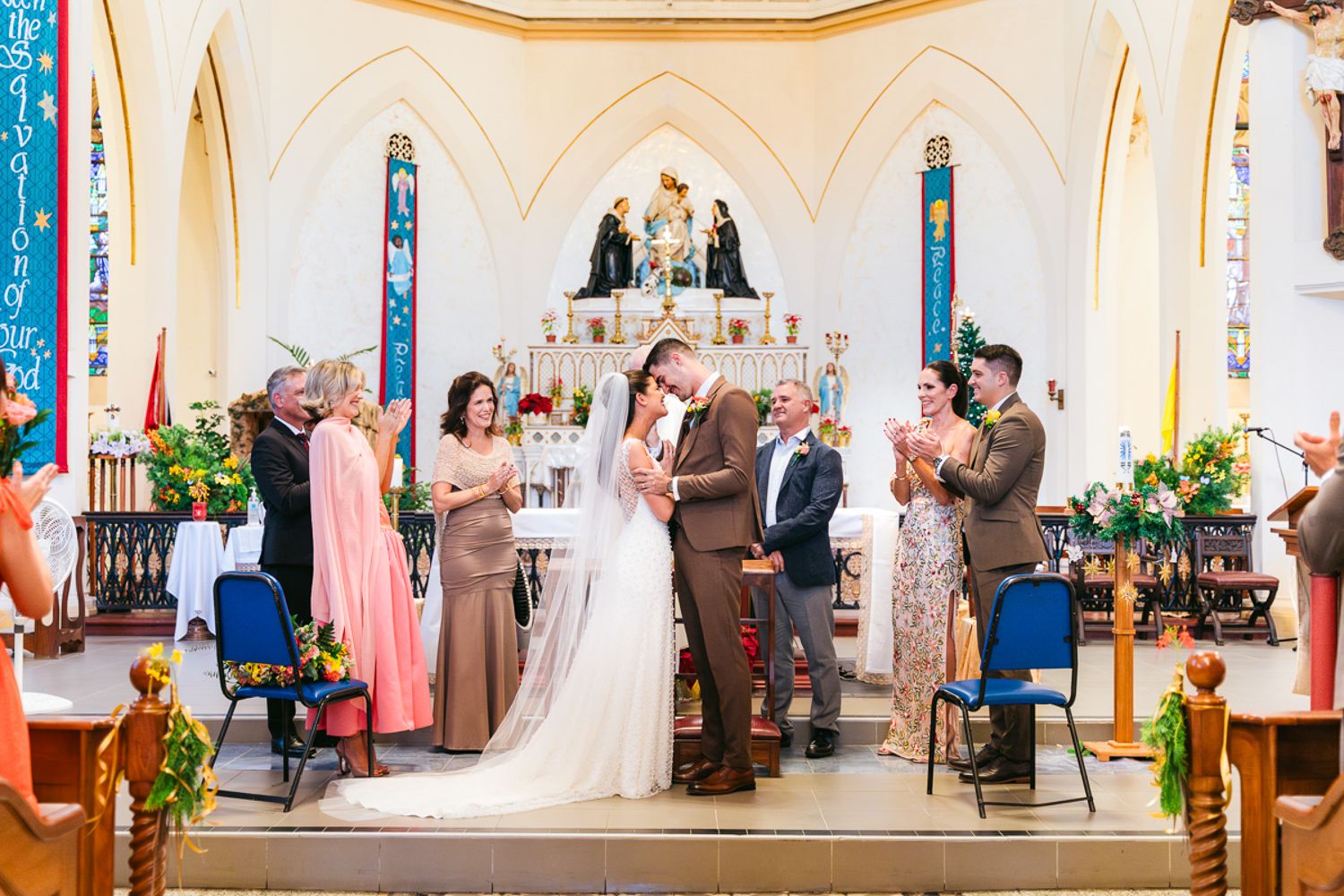 A wedding ceremony taking place in a bright white church with guests seated on wooden pews adorned with floral decorations.