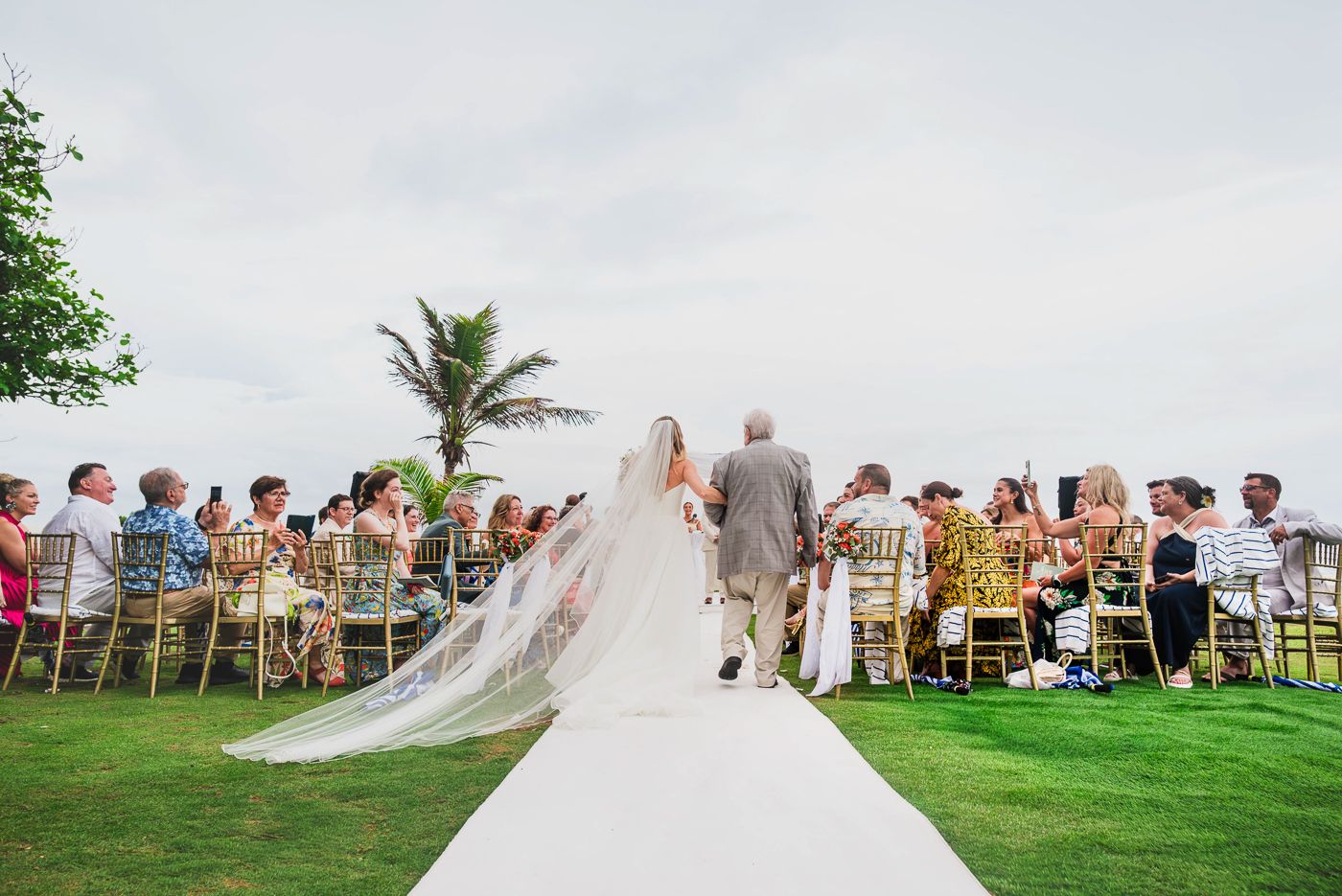 Bride walks down white aisle with long veil at outdoor beach wedding ceremony with seated guests.