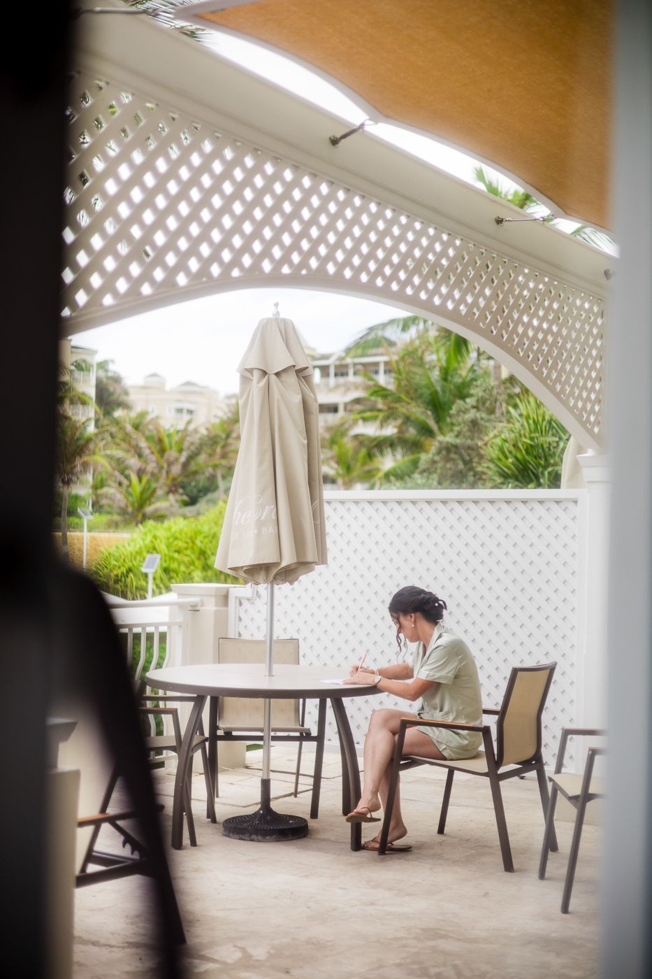 Person sitting alone at outdoor patio table under closed umbrella at tropical resort with palm trees.
