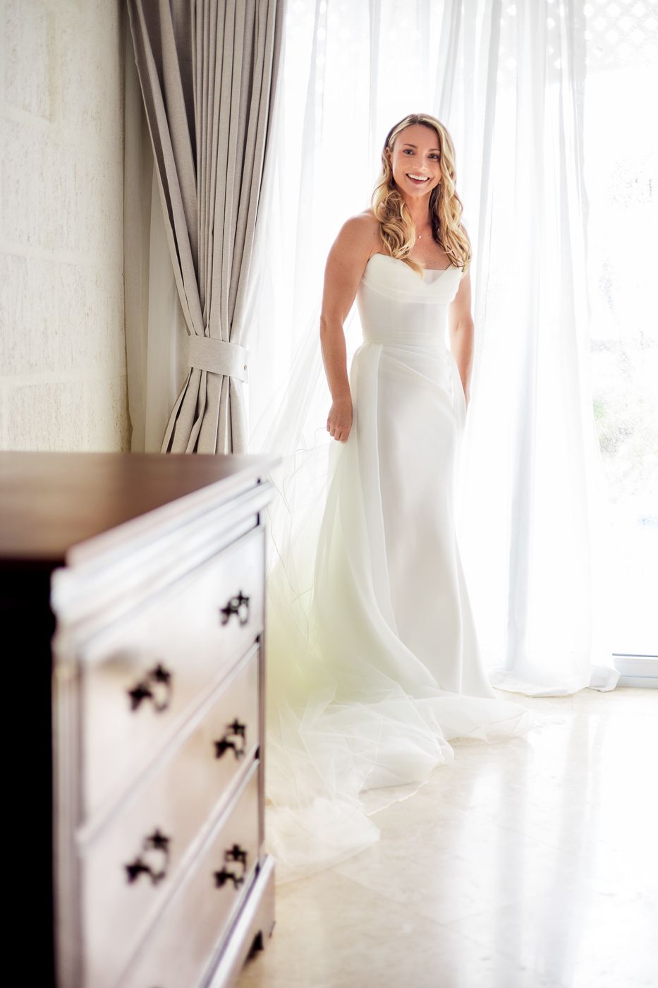 Smiling bride in a strapless white wedding gown standing by sheer curtains near a wooden dresser.