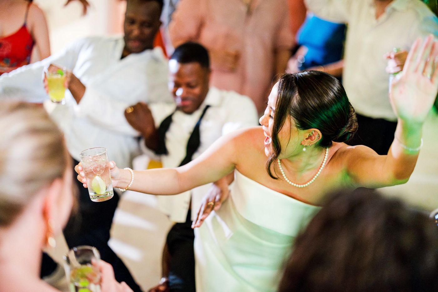 Wedding guests in light colored attire dance together at a lively reception.