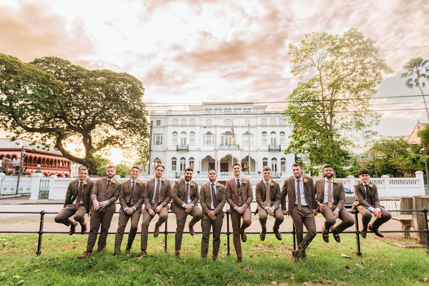 Wedding party in coordinated gray suits poses under sunset sky in a line among outdoor trees by historic building.