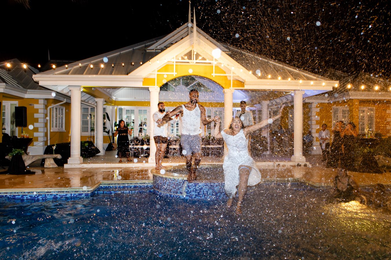 Wedding celebration with people jumping into pool while water droplets sparkle in night lighting.