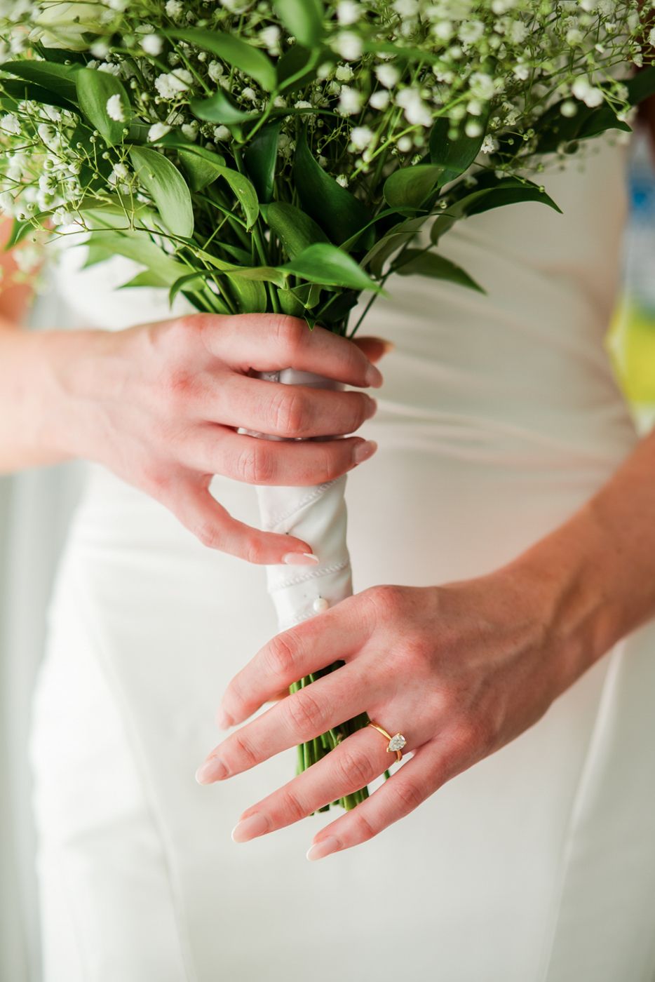 Close-up of hands holding a wedding bouquet, showing off an engagement ring and white dress details.