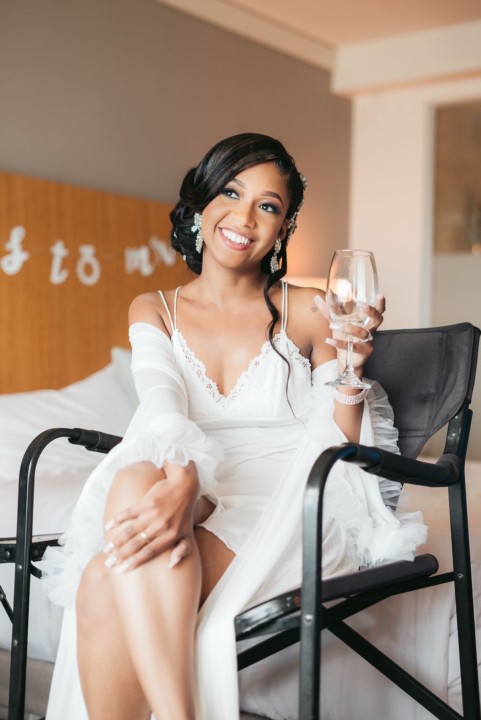 Bride in white dress sitting in chair holding champagne glass during wedding preparations.