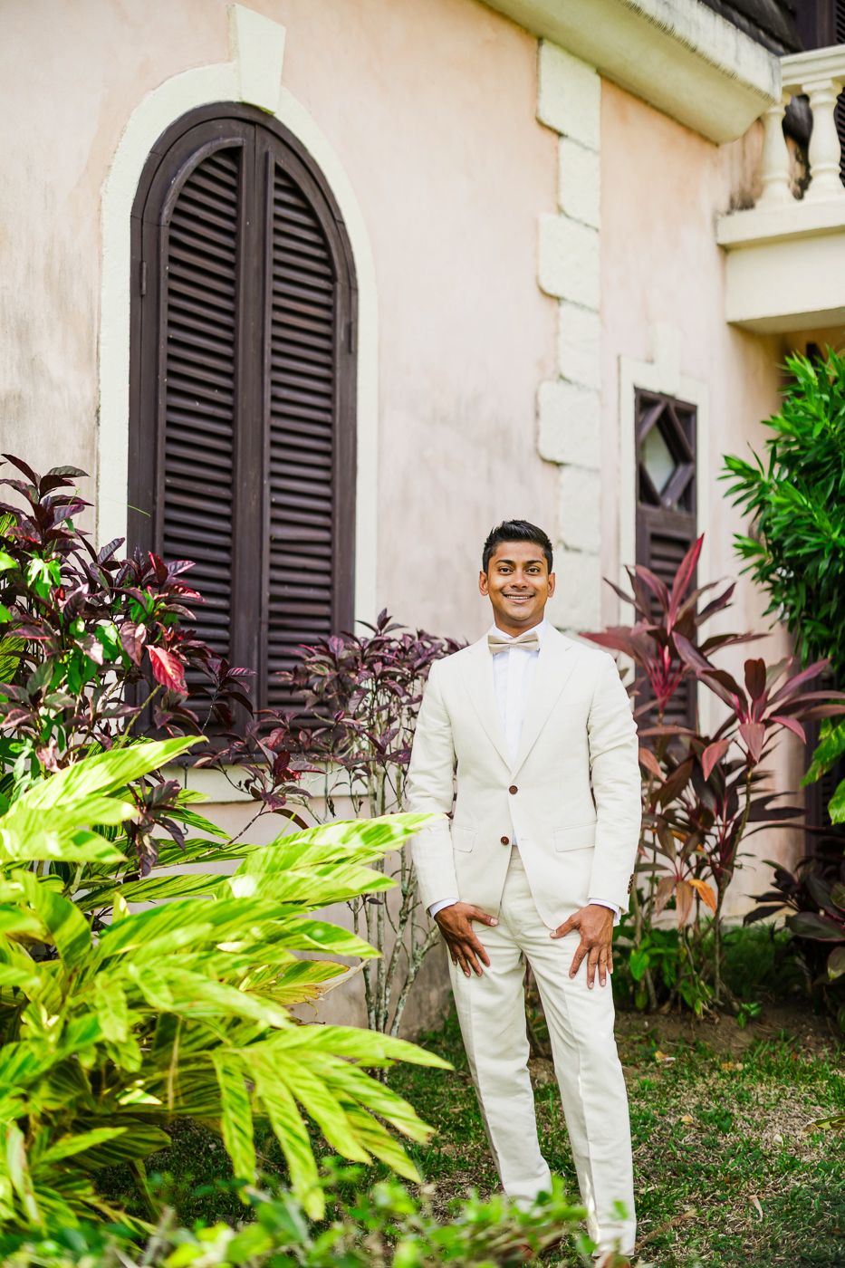 Person in all-white attire poses against tropical landscaping and shuttered windows.
