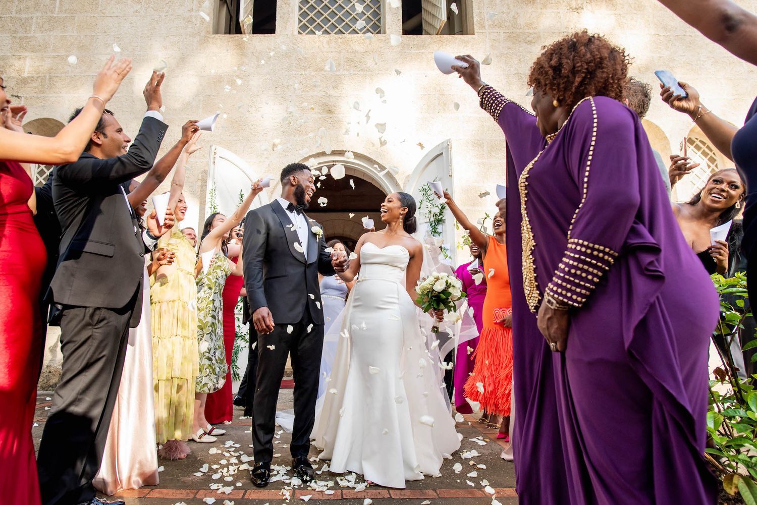Wedding guests throw flower petals during a joyful ceremony celebration outside a historic stone building.