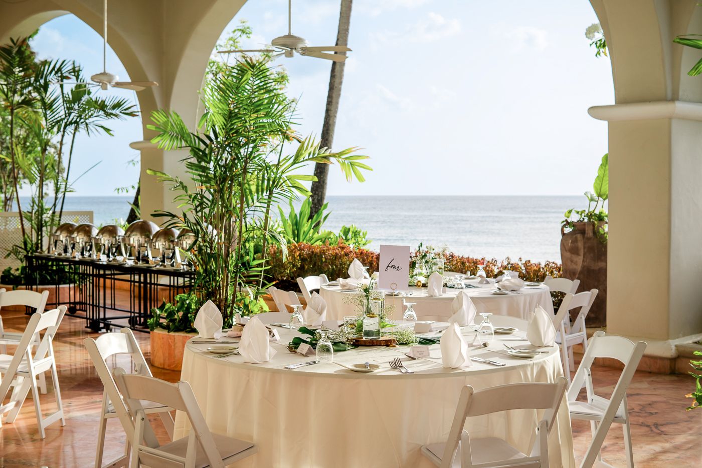 Beachfront dining setup with white tablecloths and ocean views through arched columns.