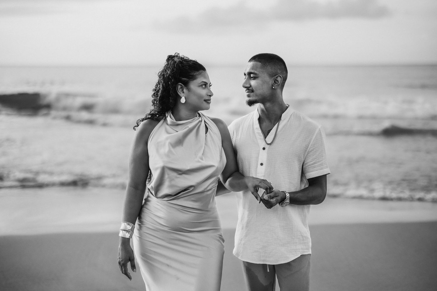 Black and white photo of a couple holding hands and gazing at each other on a beach at sunset.