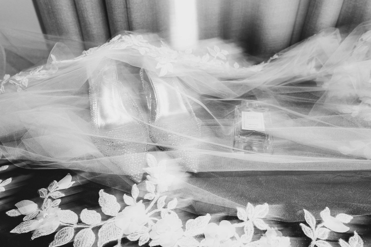 Black and white photo of a bridal veil draped over a table with lace floral details and a perfume bottle.
