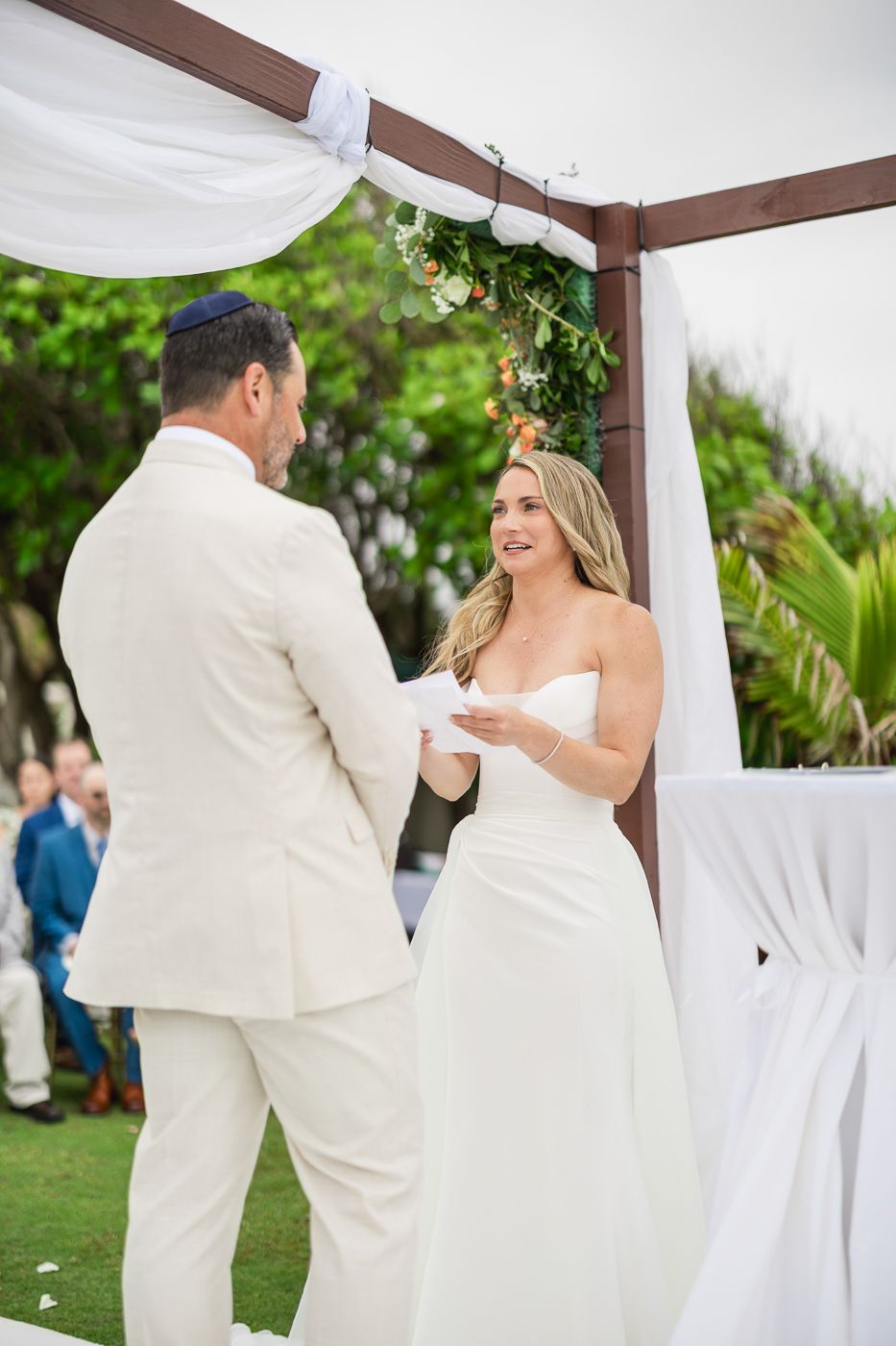 Bride in strapless white gown reads vows to groom in cream suit under floral chuppah at outdoor wedding ceremony.