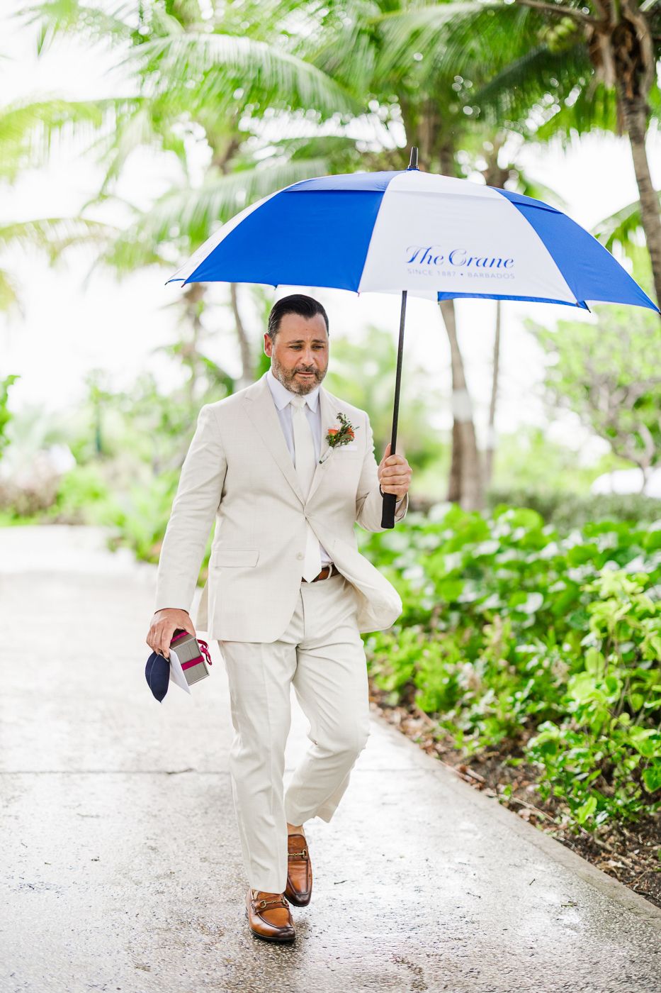 Groom in cream suit walks with blue and white The Crane umbrella on rainy wedding day at tropical venue.