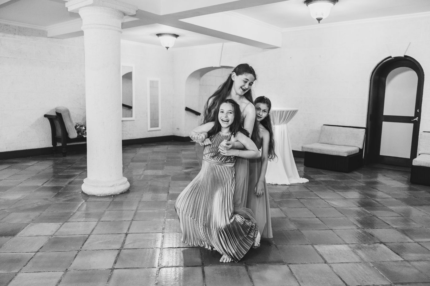 Three women in elegant dresses pose together in a black and white photo inside a grand hall.