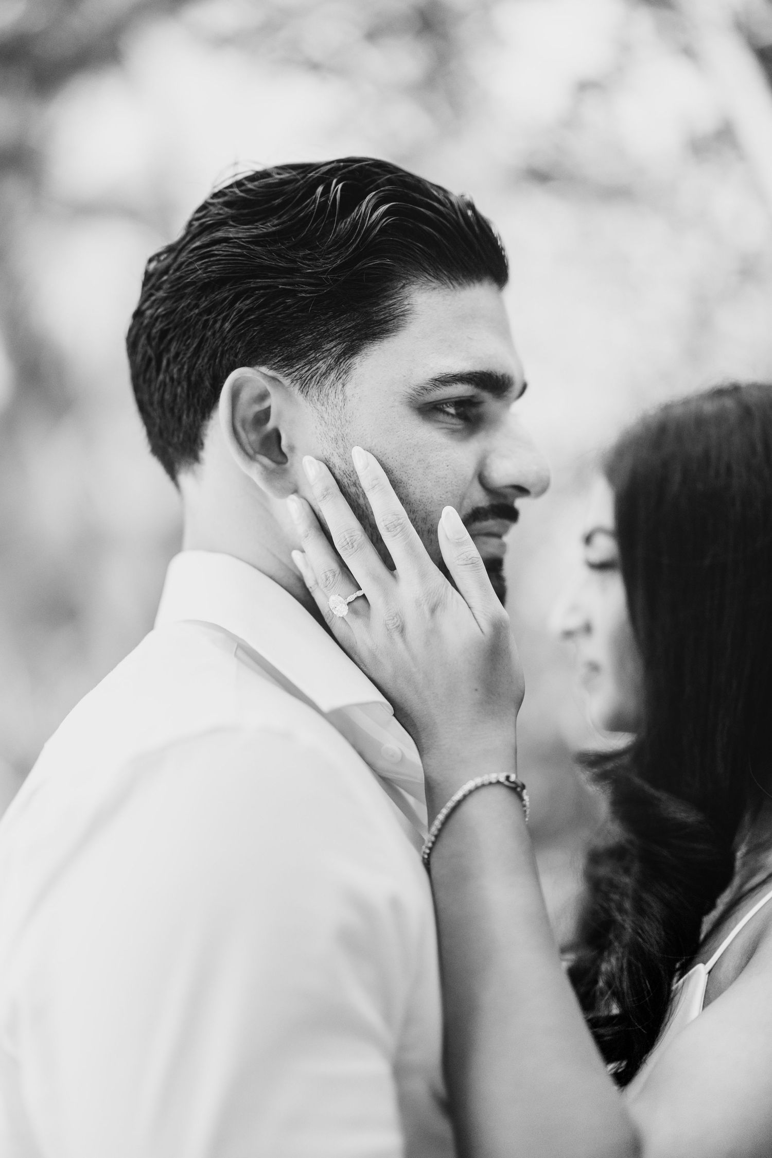 Black and white engagement photo of a woman gently touching her fiancé's cheek, her diamond ring and bracelet visible.
