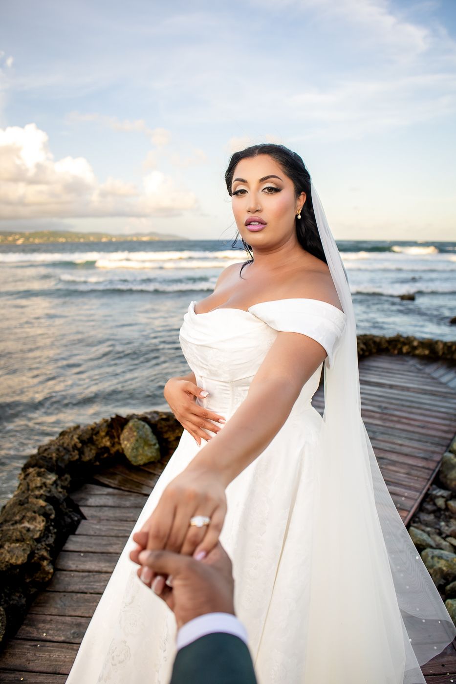 Bride in off-shoulder white gown holds groom's hand on ocean pier at sunset.