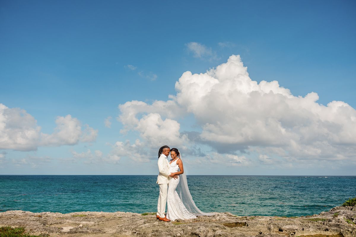 Bride and groom embrace on rocky shore with turquoise ocean and blue sky backdrop at destination wedding.