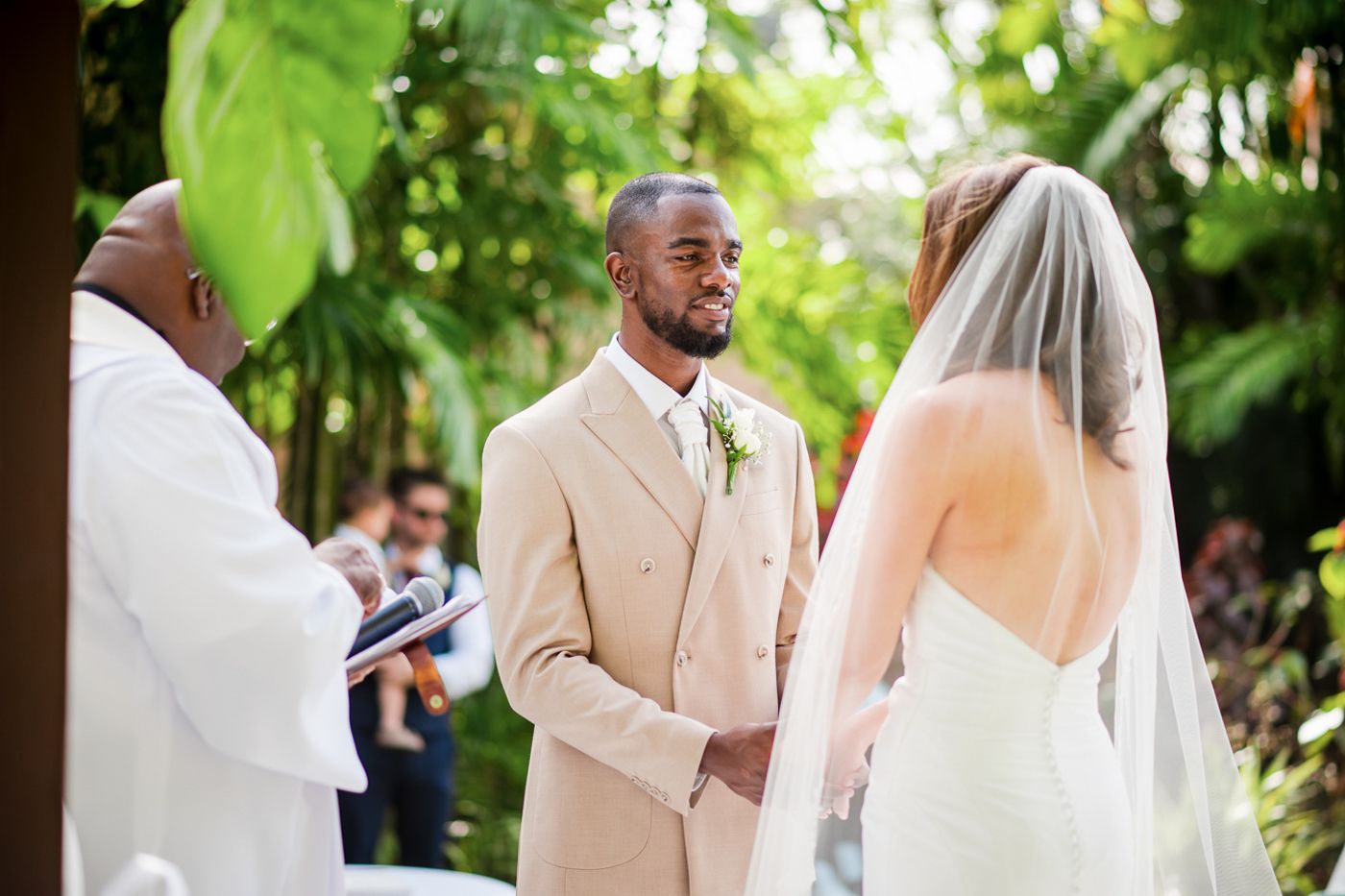 Wedding ceremony taking place under tropical foliage with white floral arrangements.