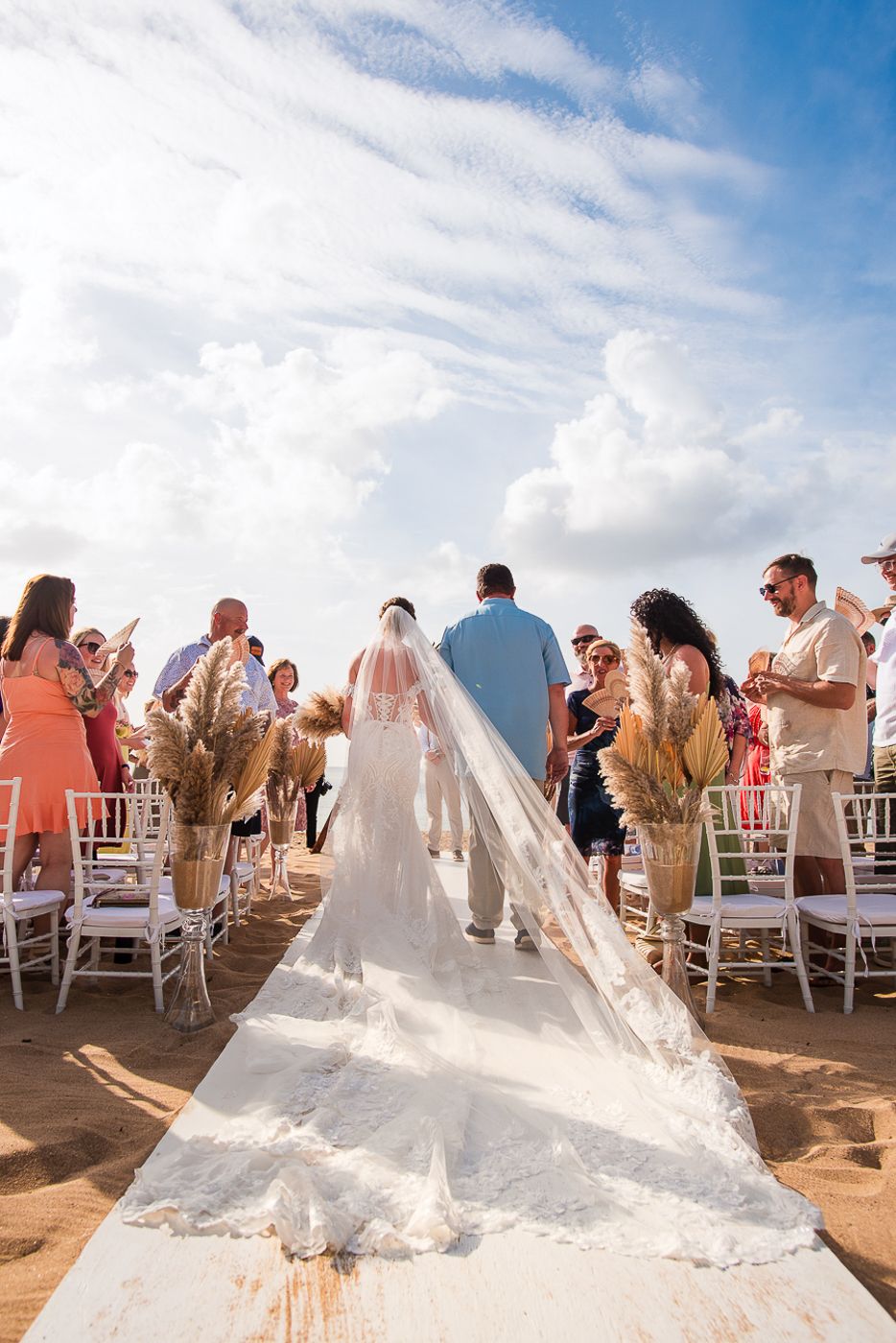 Wedding ceremony on a beach with flowing white dress train and dried pampas grass bouquets.