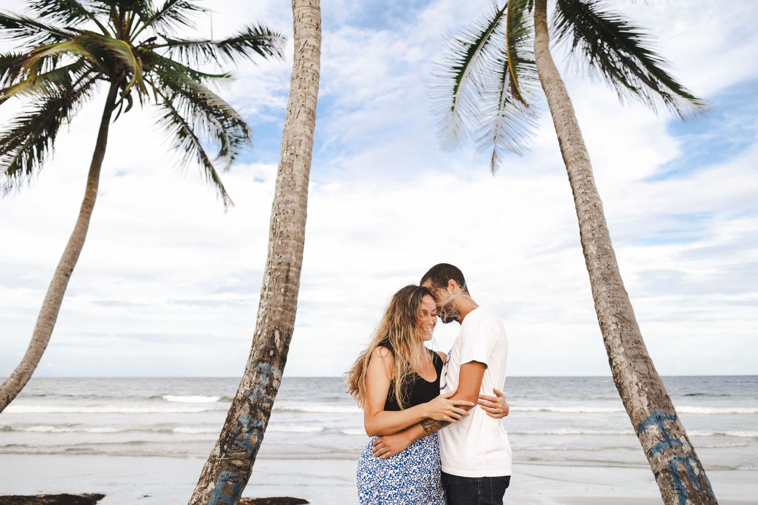 Couple embraces between palm trees on tropical beach during sunset.