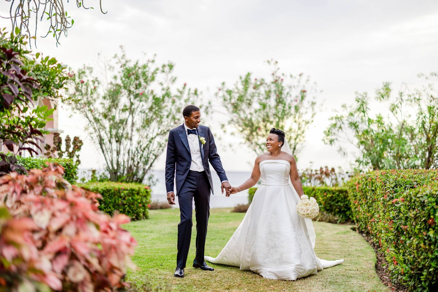 Bride and groom hold hands walking through lush garden path on their wedding day, wearing elegant white gown and navy tuxedo.
