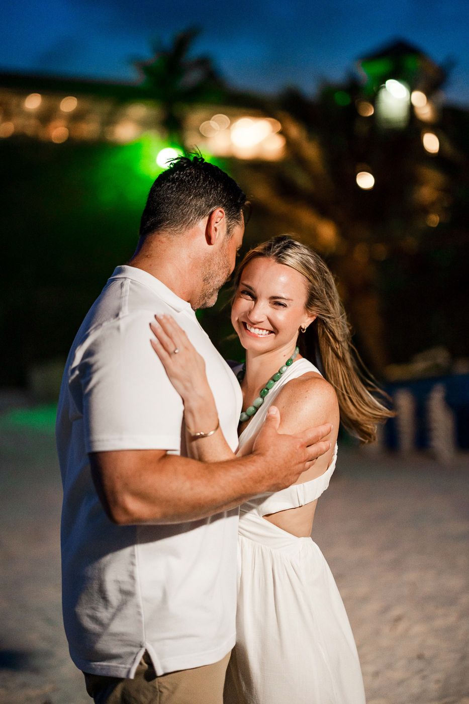Couple dancing and embracing on a beach at night, woman smiling, both wearing white outfits with tropical resort lights behind.