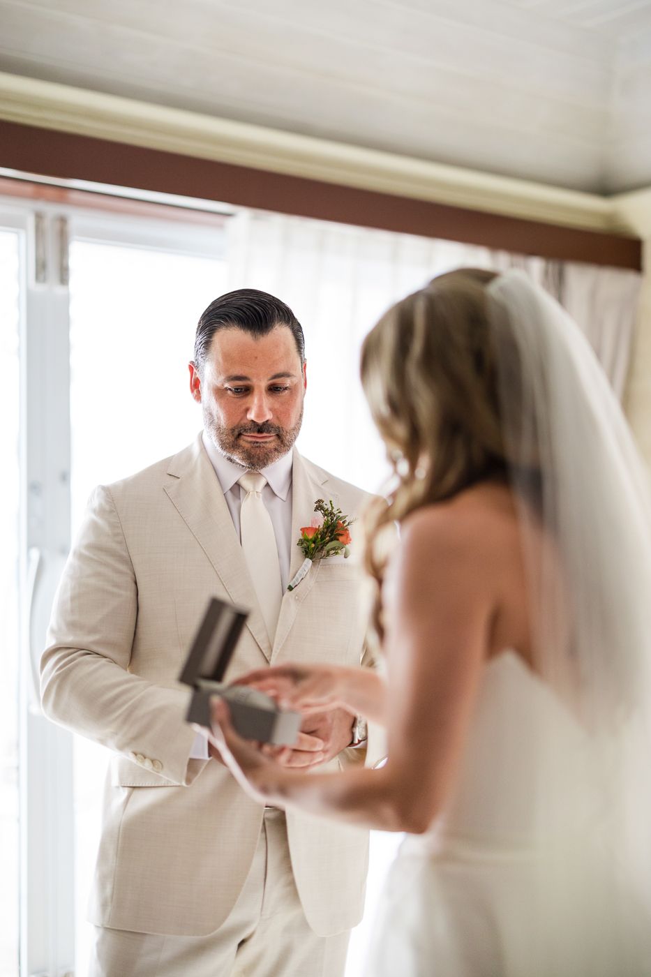 Groom in ivory suit opens ring box during wedding ceremony while bride in white gown and veil stands before him.