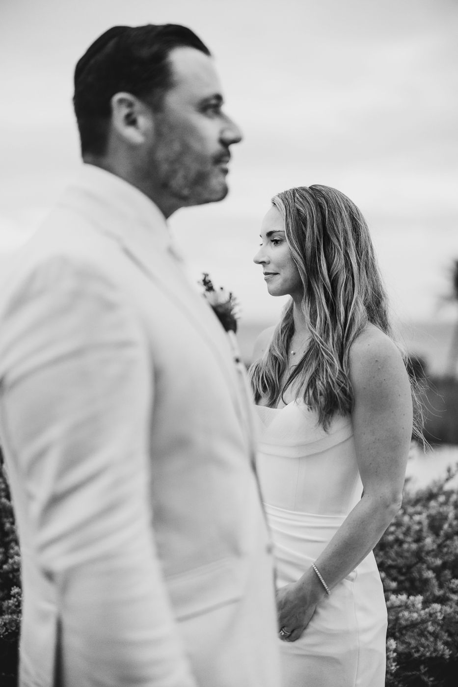 Black and white wedding photo of bride in white dress gazing at groom in white suit outdoors.