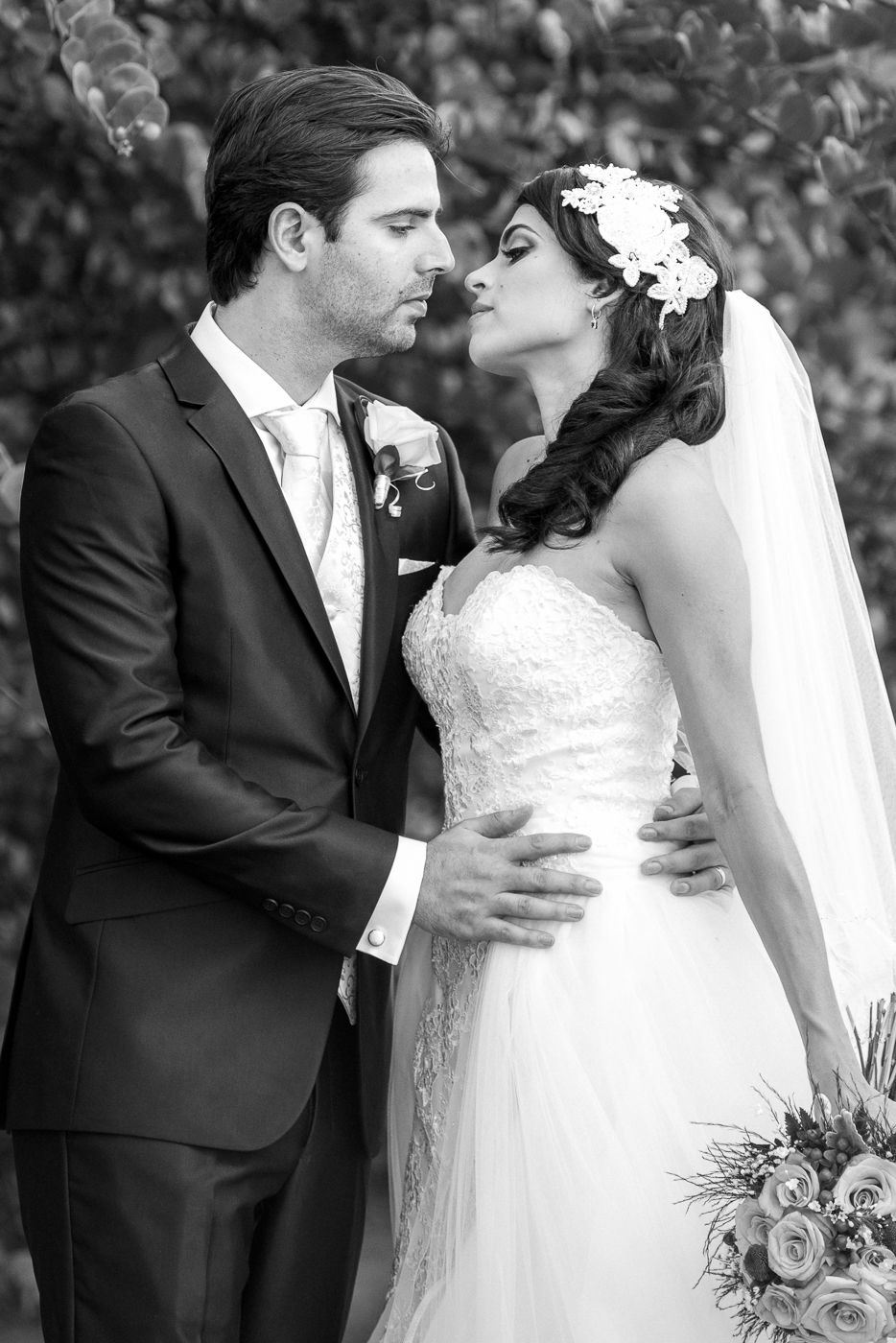 Black and white wedding photo of bride and groom sharing a romantic moment outdoors, bride in lace gown with floral headpiece.