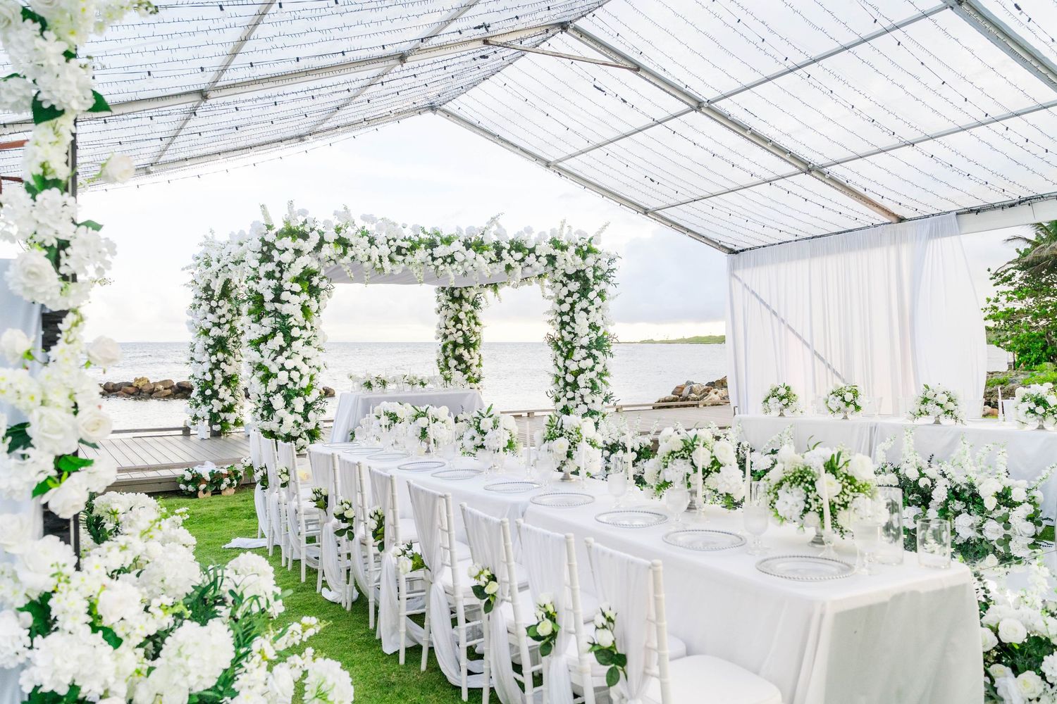 Elegant all-white wedding reception under a clear tent with floral arches, long dining table, and lush white flower arrangements.