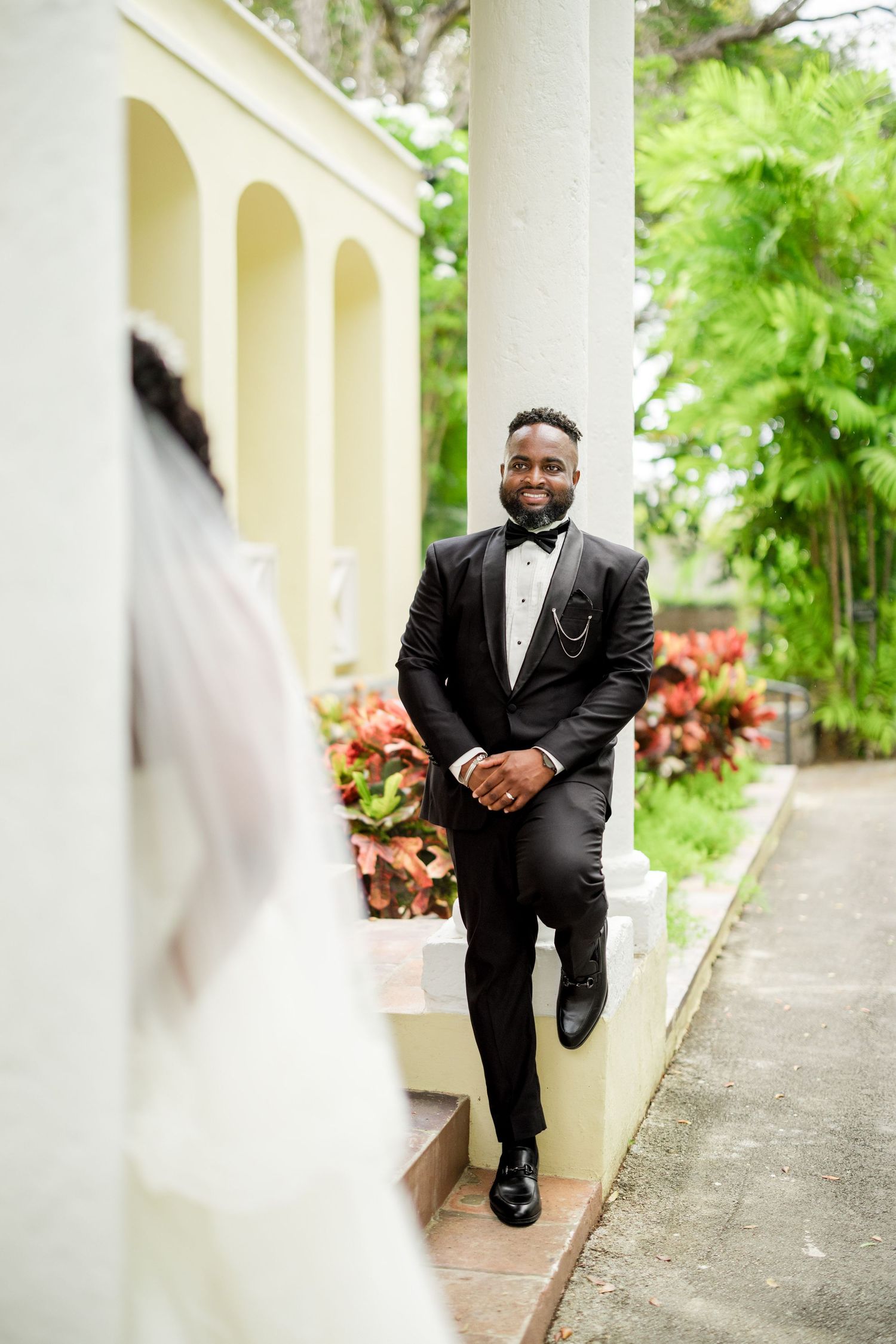 Groom in black tuxedo smiling at bride during outdoor wedding ceremony near elegant columns and tropical greenery.