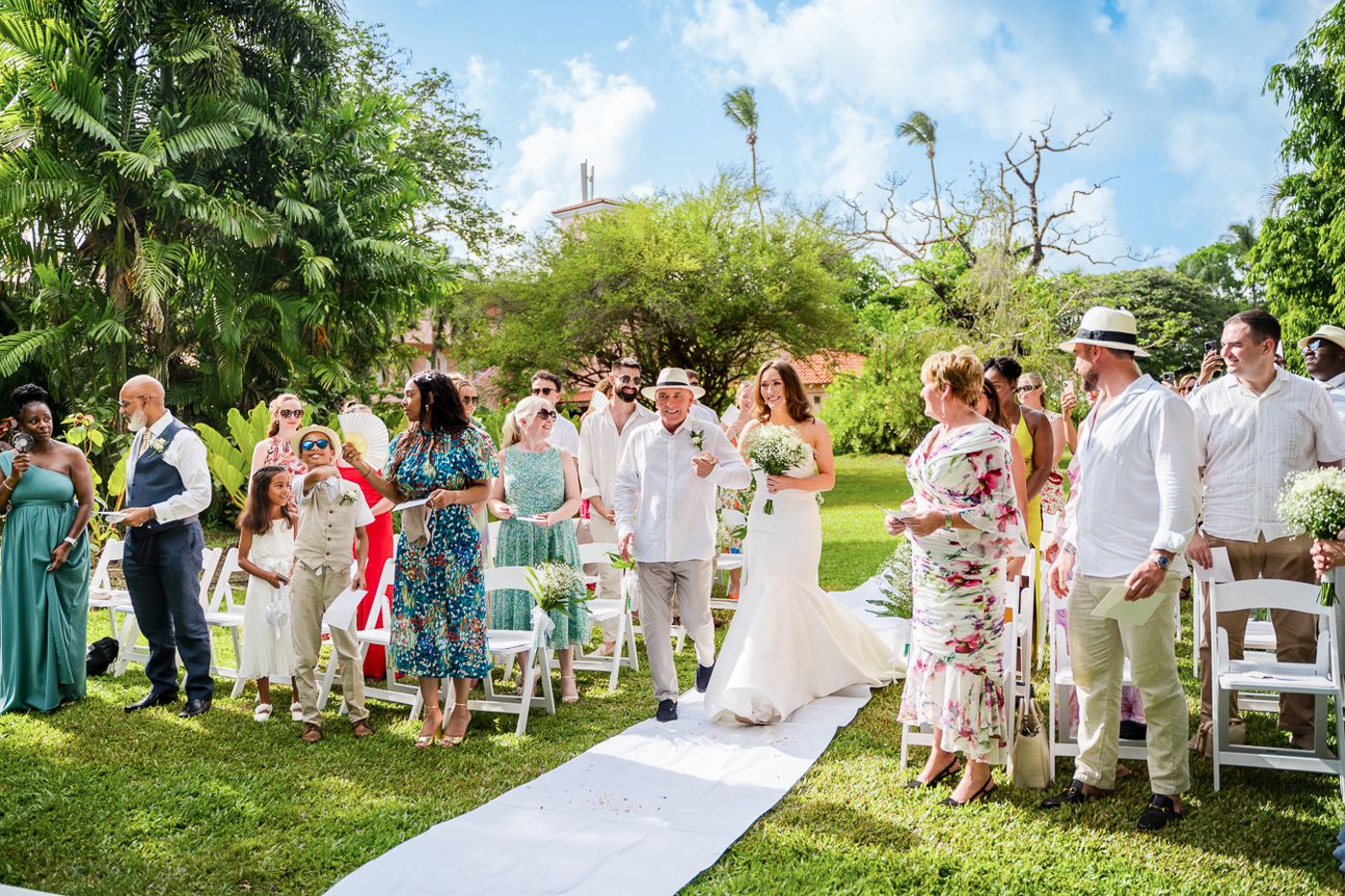 Wedding guests gather on a lush green lawn with palm trees for an outdoor tropical ceremony.