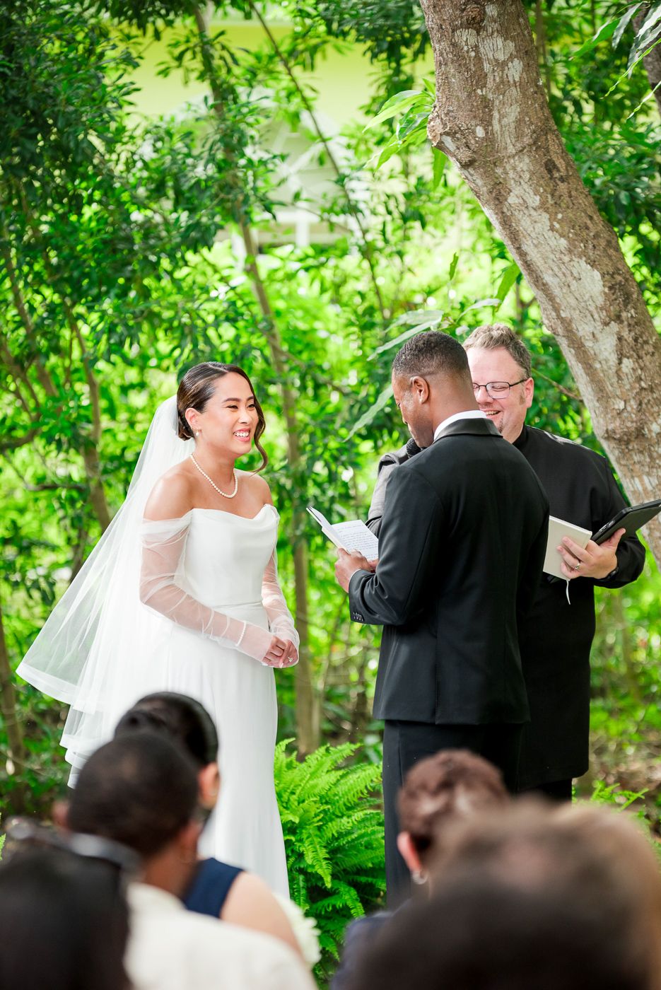 A wedding ceremony takes place on a white aisle runner under tropical trees with guests seated on both sides.