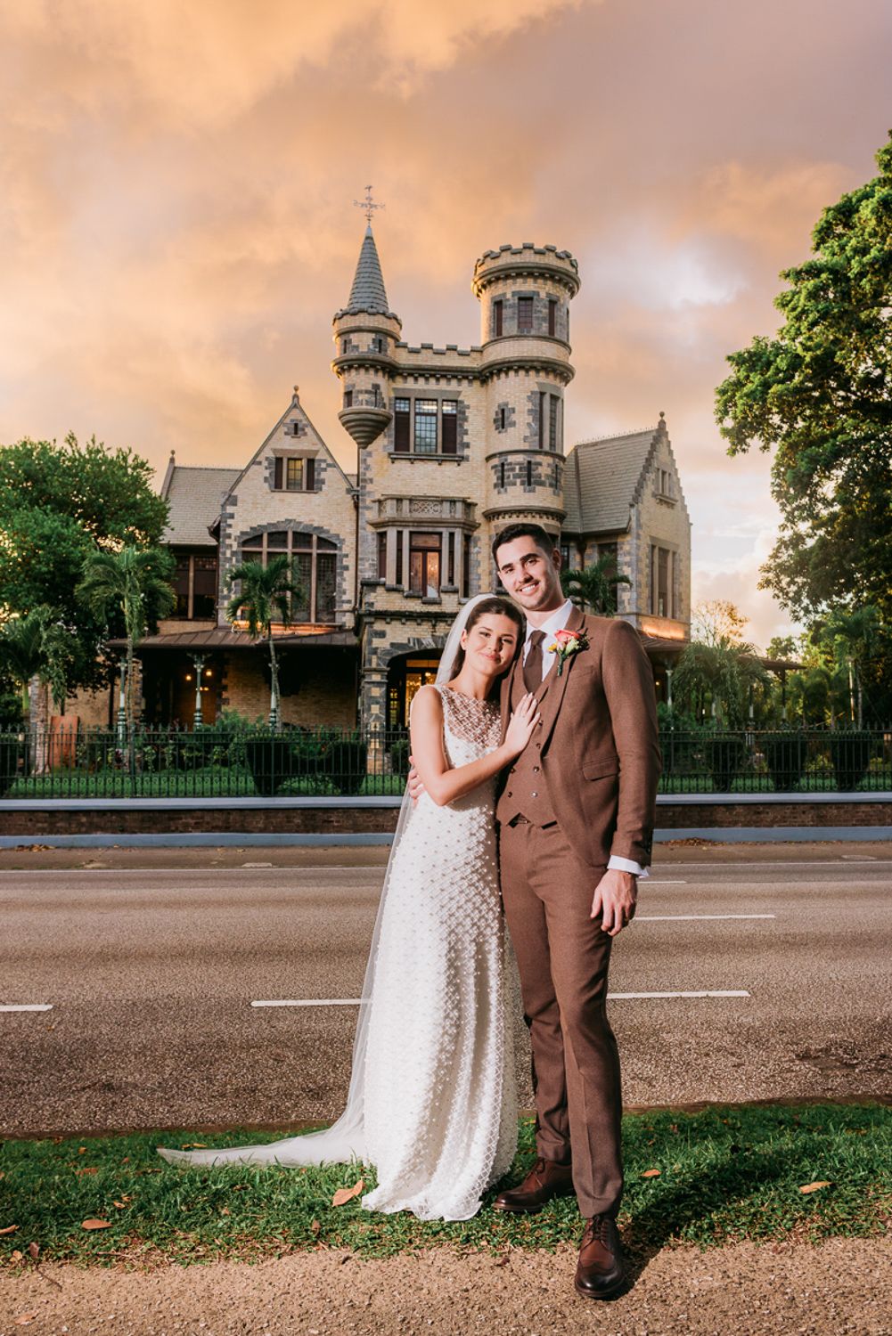 A couple stands together in formal attire at sunset in front of a historic Victorian-style church building.