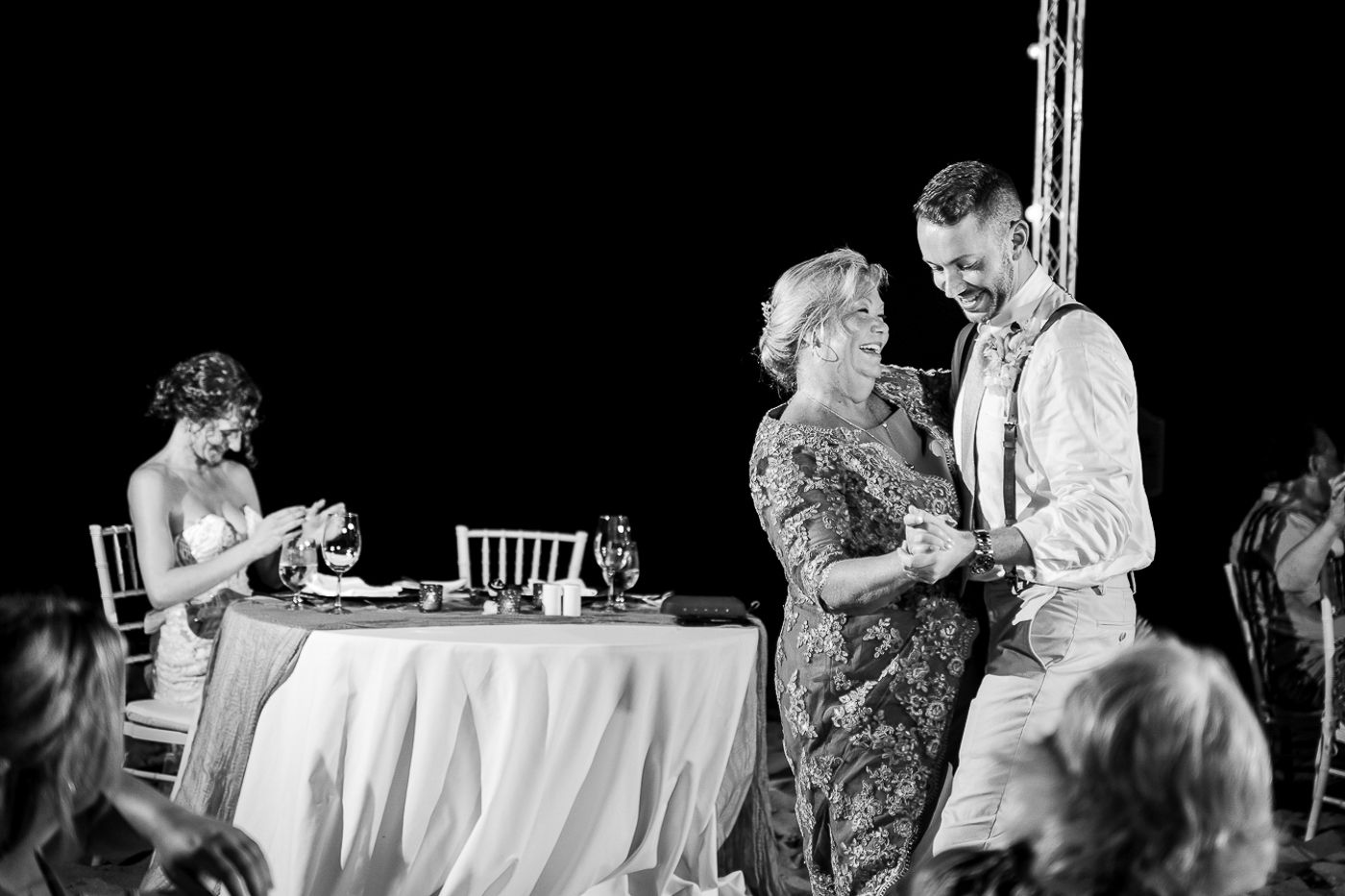 Wedding party guests share laughs and conversations at a beach reception under twinkling string lights.
