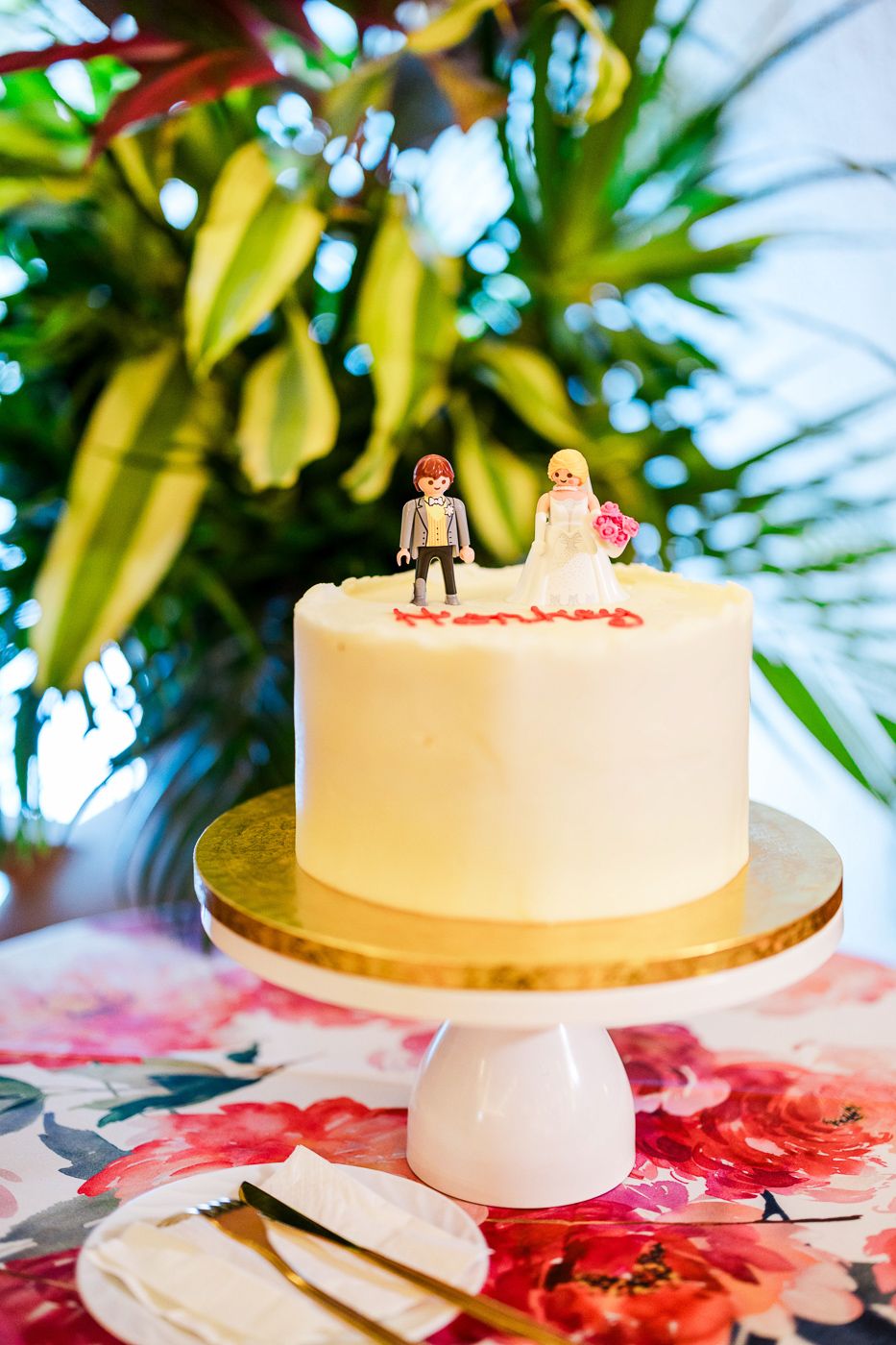 Simple white wedding cake with figurine toppers displayed against tropical foliage.