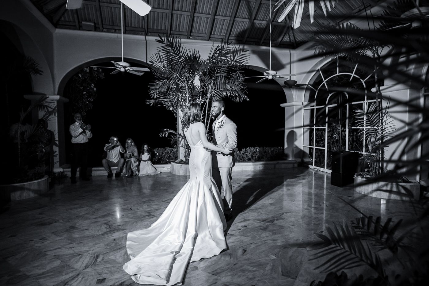 Black and white photo of a dancing couple in formal attire under ornate arches with tropical plants.