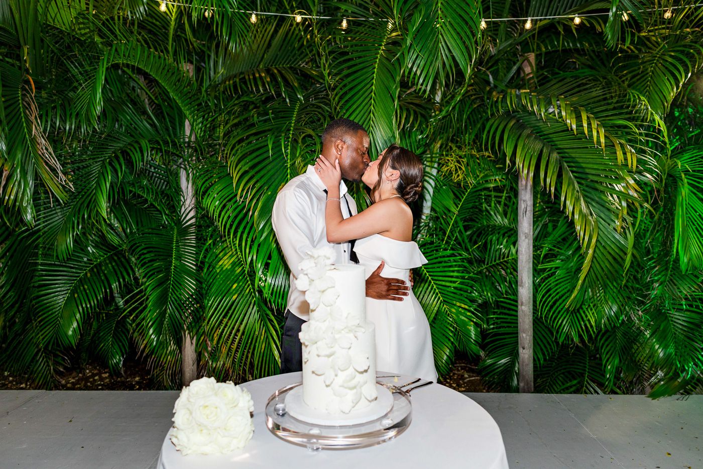 Wedding guests dancing and celebrating at a tropical outdoor reception with palm trees and string lights.