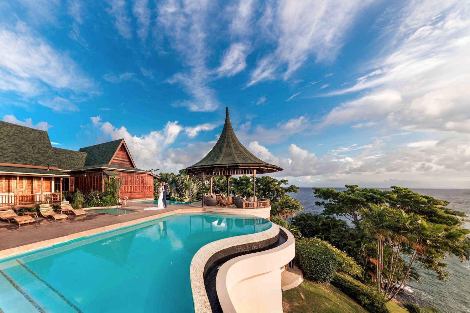 Luxury tropical villa with infinity pool overlooking ocean, featuring elegant gazebo and lush palm trees under blue sky.