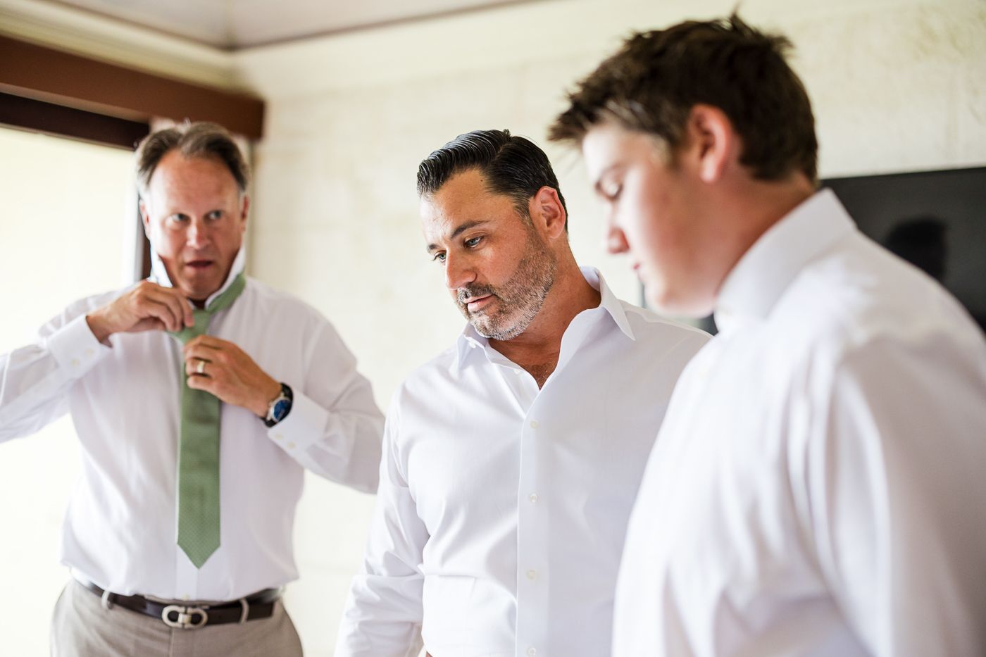 Three men in white dress shirts preparing for a wedding, with one helping adjust a green tie.