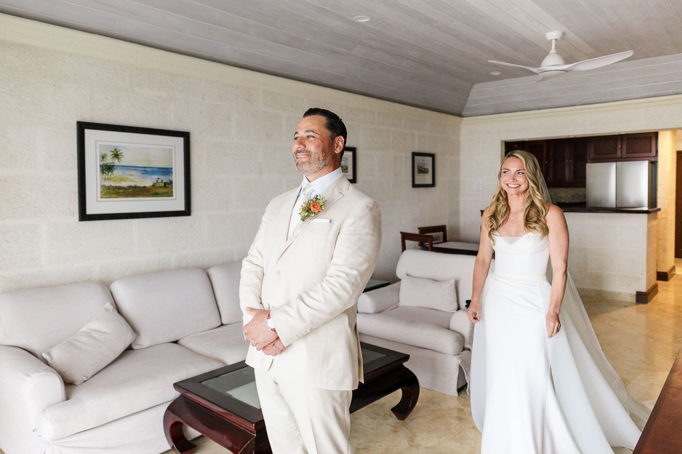 Bride in white gown stands behind groom in cream suit during first look moment in elegant resort suite.
