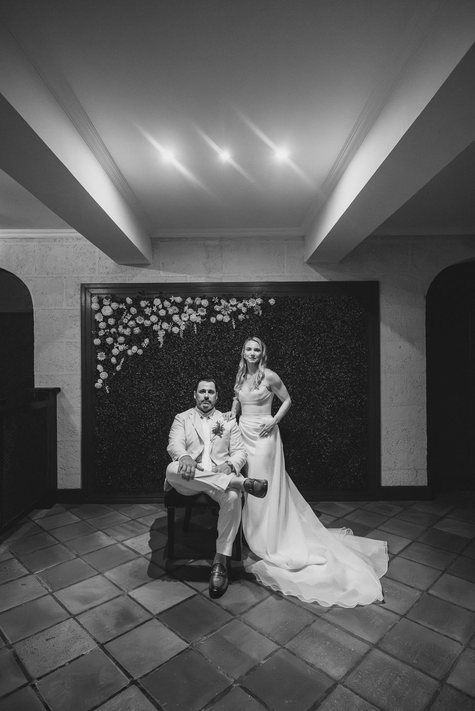 Black and white wedding portrait of bride and groom posing before a floral wall backdrop in an elegant indoor venue.