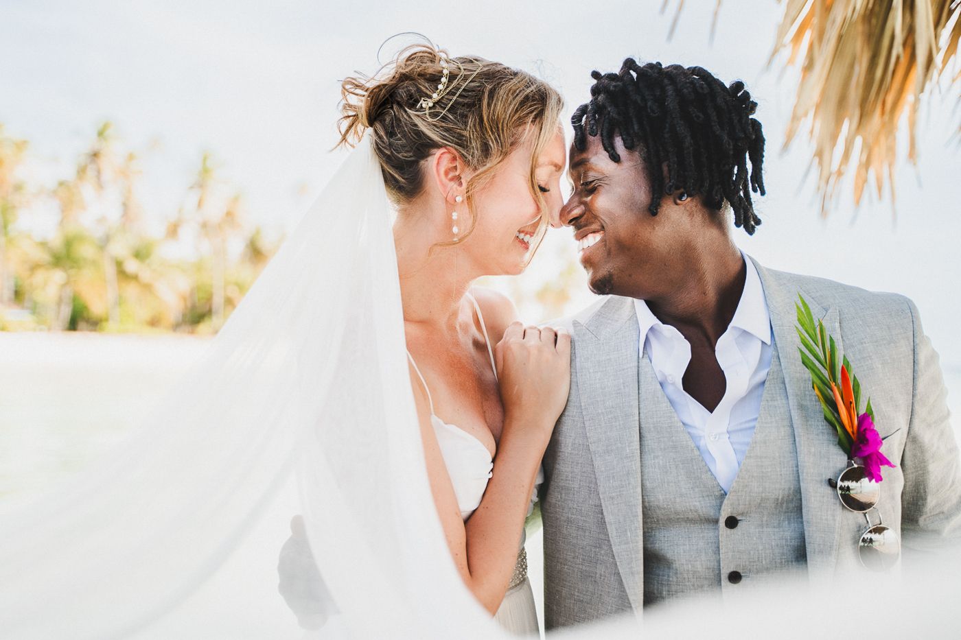 Interracial couple sharing a romantic moment at a tropical beach wedding, bride in veil and groom in gray suit.