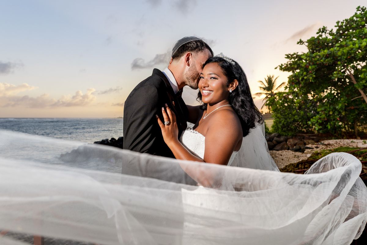 A couple shares a romantic moment on a tropical beach as a flowing white veil catches the ocean breeze at sunset.