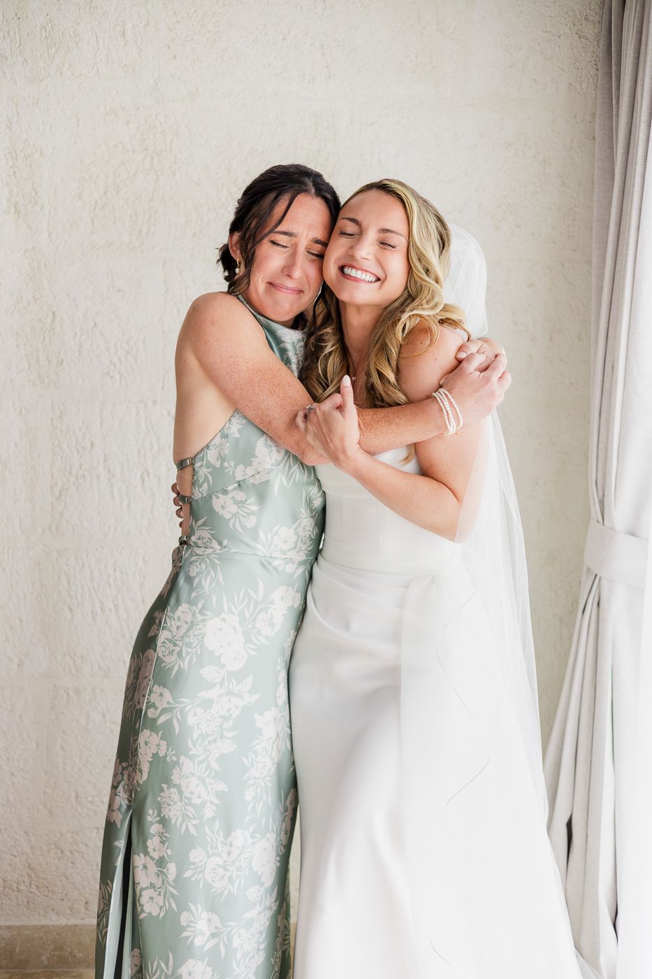 Bride in white gown and veil hugs smiling woman in sage floral dress during wedding day preparations.