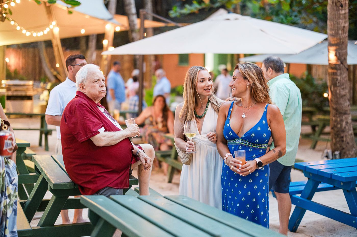 People socializing outdoors at a tropical beach bar, enjoying drinks at green picnic tables with string lights.